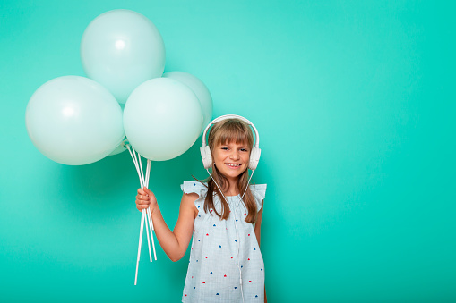 Portrait of a beautiful little girl holding bunch of balloons wearing headset and listening to the music, isolated on mint colored background with copy space