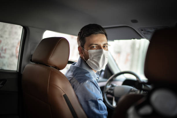 Driver taking to a passenger on seat back wearing protective medical mask Driver taking to a passenger on seat back wearing protective medical mask mid adult men stock pictures, royalty-free photos & images