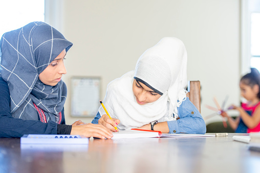 A Muslim mother and her young daughter sit at a table in casual clothing and wearing Hijabs. The mother is helping her daughter with her writing homework and they are both looking down at the notebook.