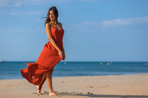 Pretty young woman in a red dress and nude foots on the sand of the beach. Happy girl enjoying freedom at the sea.