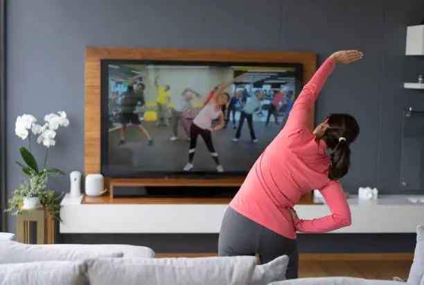 Back view of senior woman following an online stretching class looking at TV screen - Technology concepts. **IMAGE ON SCREEN BELONGS TO US**