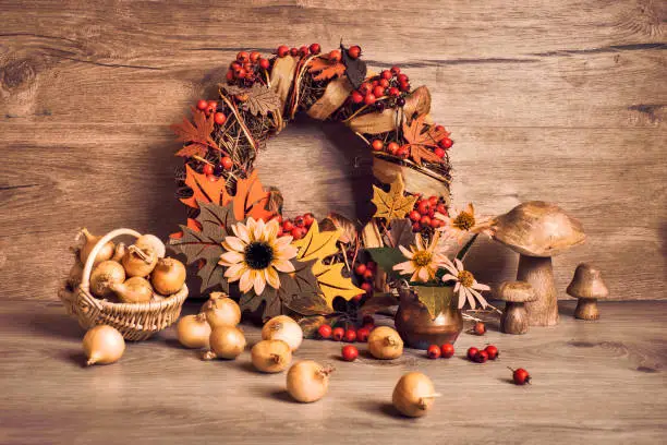 Autumn Thanksgiving arrangement with wreath decorated with orange berry felt Fall leaves and ribbons. Basket of onions, decorative pumpkins, Autumn leaves and flowers on aged wooden table, mushrooms.