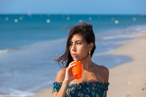 Portrait of a beautiful brunette drinking cocktail while standing on beach. Summer vacation concept. Happy girl enjoying freedom.