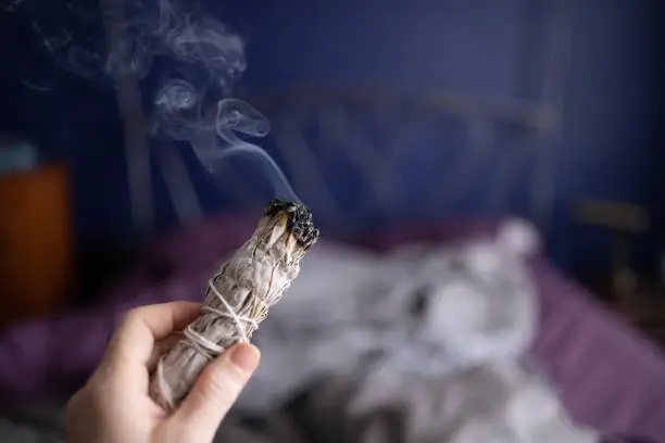 This is a photograph of a burning bundle of sage as a woman’s hand smudges indoors during the shelter in place order during the coronavirus pandemic in Miami, Florida USA.