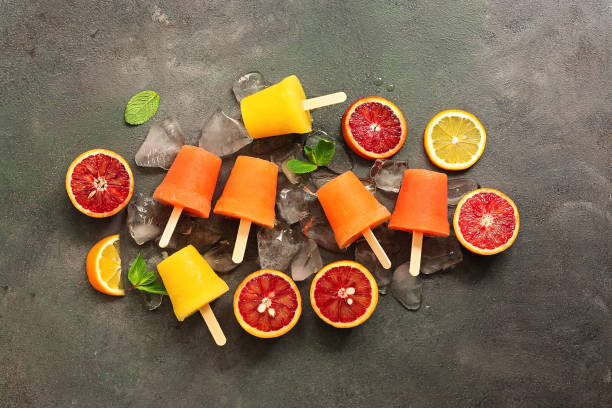 istock Assorted orange popsicles with slices of citrus, mint and ice on a dark rustic background. Frozen juice of red and yellow orange on a stick. Top view, flat lay. 1216722135