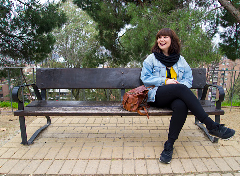 Portrait of a urban young stylish smiling woman in bright modern clothes sitting on a bench. She is looking at the side. Lifestyle and urban concept.