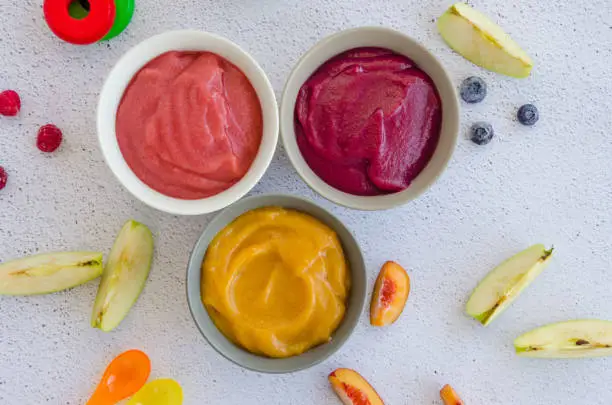 Baby food. Homemade fruit puree. Variety of apple puree or applesauce with frozen peach, raspberries and blueberries in three bowls on a light background. Healthy food. Horizontal. Top view