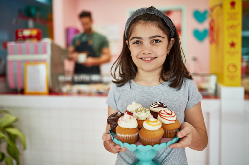Smiling 9 year old Hispanic girl holding dessert stand filled with gourmet cupcakes in Miami vegan bakery.