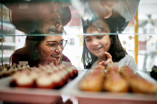 Focus on background of Hispanic mother and 9 year old daughter smiling as they pick gourmet cupcakes from bakery display case.