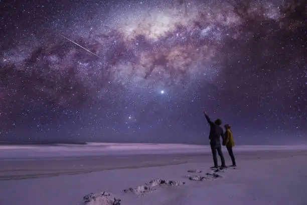 Couple holding hands looking and pointing in amazement at a shooting star while standing on a secluded beach.