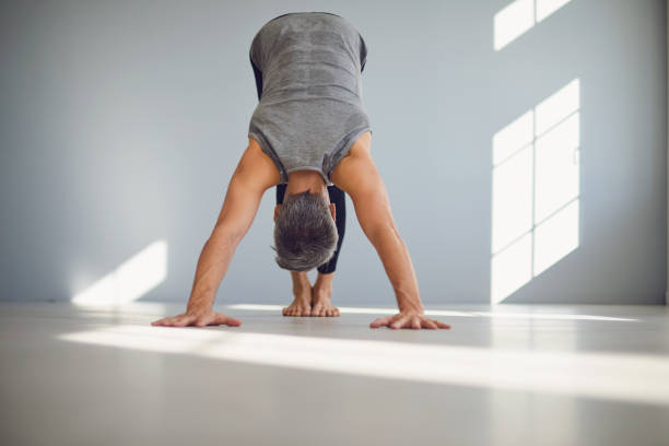 Gray haired flexible man practicing yoga in dog pose in light modern studio Low angle of barefooted unrecognizable male in sportswear focusing and doing downward facing dog yoga exercise while training alone at home grey hair on floor stock pictures, royalty-free photos & images