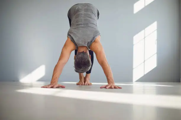 Photo of Gray haired flexible man practicing yoga in dog pose in light modern studio