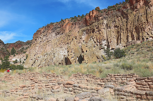 Part of the Tyuonyi ruins of the ancestral Pueblo peoples an unidentifiable hikers by the cliffs along the main loop trail in Frijoles Canyon at Bandelier National Monument near Los Alamos, New Mexico against a bright blue autumn sky