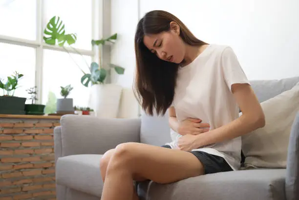 Photo of Asian girl having period is sitting on sofa and feeling much of painful on her stomach with unknown reason. She holds onto her stomach and curls through spasms. Healthcare and medical concept.