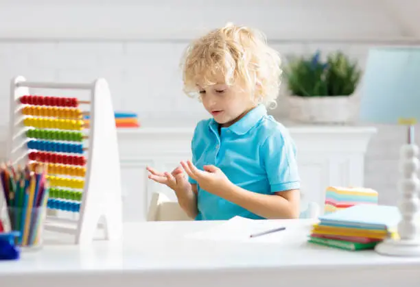 Child doing homework at home. Little boy with wooden colorful abacus doing math exercise learning addition and counting. Kids study and learn. Preschooler kid writing and reading. Back to school.