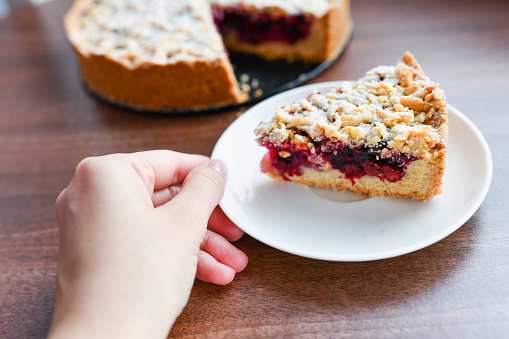Pie with berries: raspberries, strawberries, currants, on a white plate,a woman's hand holding a piece of cake on a spatula. On a wooden background, in the background linen napkin.