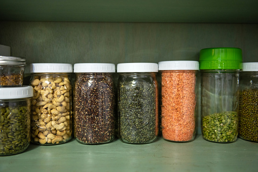 This is a candid photograph of a kitchen pantry shelf lined with jars full of vegan dry food including quinoa, lentils, green peas, and beans at home.