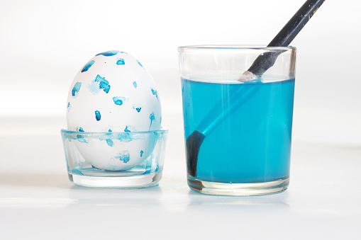 Easter egg painted with blue dots and a glass with a brush and blue colored water forhomemade holiday decoration, gray background with copy space, selected focus