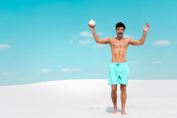 glücklicher mann mit muskulösen oberkörper in badeshorts mit kokosgetränk am sandstrand - swim truncks stock-fotos und bilder