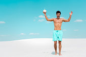 happy man with muscular torso in swim shorts with coconut drink on sandy beach
