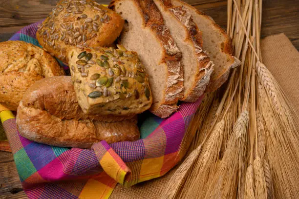 Freshly various buns and bread slices in a basket and a half whole grain bread on a jute fabric placed on a rustic wooden background with grain ears.