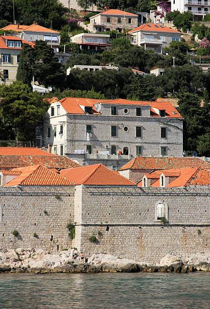 Aerial view of a coastal town featuring historic stone buildings along the rocky shoreline and turquoise sea. Scenic Mediterranean hillside with dense housing and a marina filled with boats. Seaside travel destination.