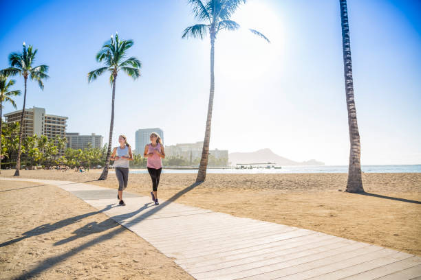 zwei frauen joggen auf der strandpromenade zwischen zwei palmen - women hawaii islands beach beauty in nature stock-fotos und bilder