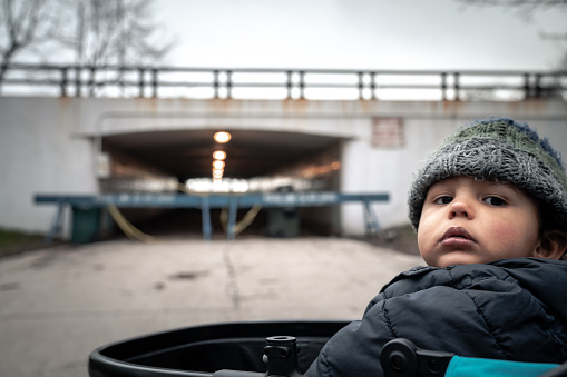 Young baby boy sits in stroller looking at camera wearing knit hat and jacket with police barricades blocking an underpass leading to lakefront in background in Chicago during the COVID-19 outbreak.