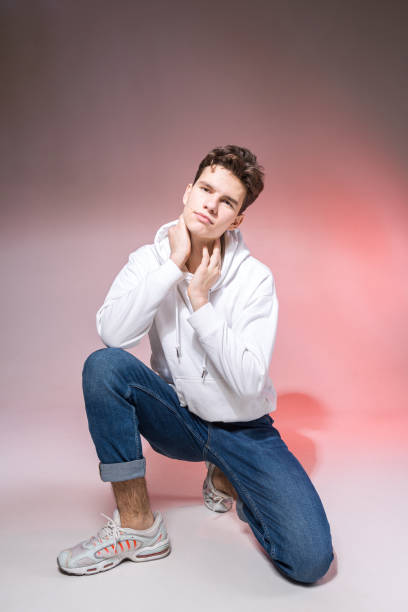 istock Handsome young stylish man sitting on the floor in studio. Teenager model posing while sitting on a pink background in casual clothes. The guy in jeans, sneakers and a white jacket with a hood 1216851097