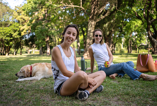 Two gorgeous female friends sitting and relaxing in a public park with their golden labrador dog