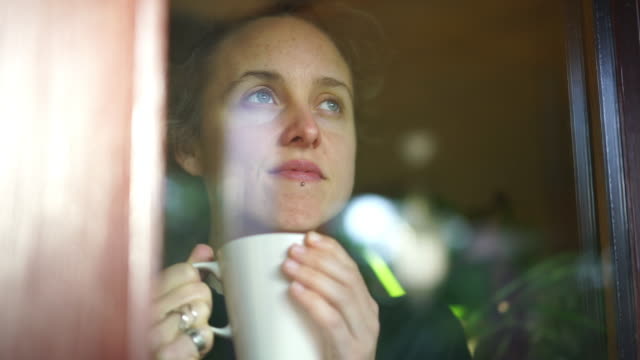 Woman drinking hot beverage looks out of window from inside her apartment