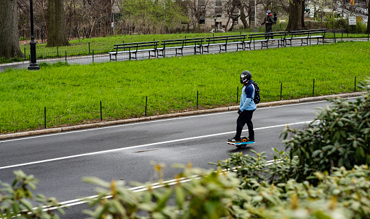 NewYork, NY, USA - April 4, 2020: A  man rides north on the East Drive of New York’s Central Park using his electric skateboard, made by OneWheel. In the background a woman wears a protective mask due to the ongoing coronavirus pandemic.