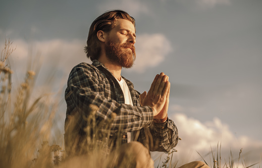 From below side view of adult bearded male in casual checkered shirt sitting in lotus pose in field and meditating