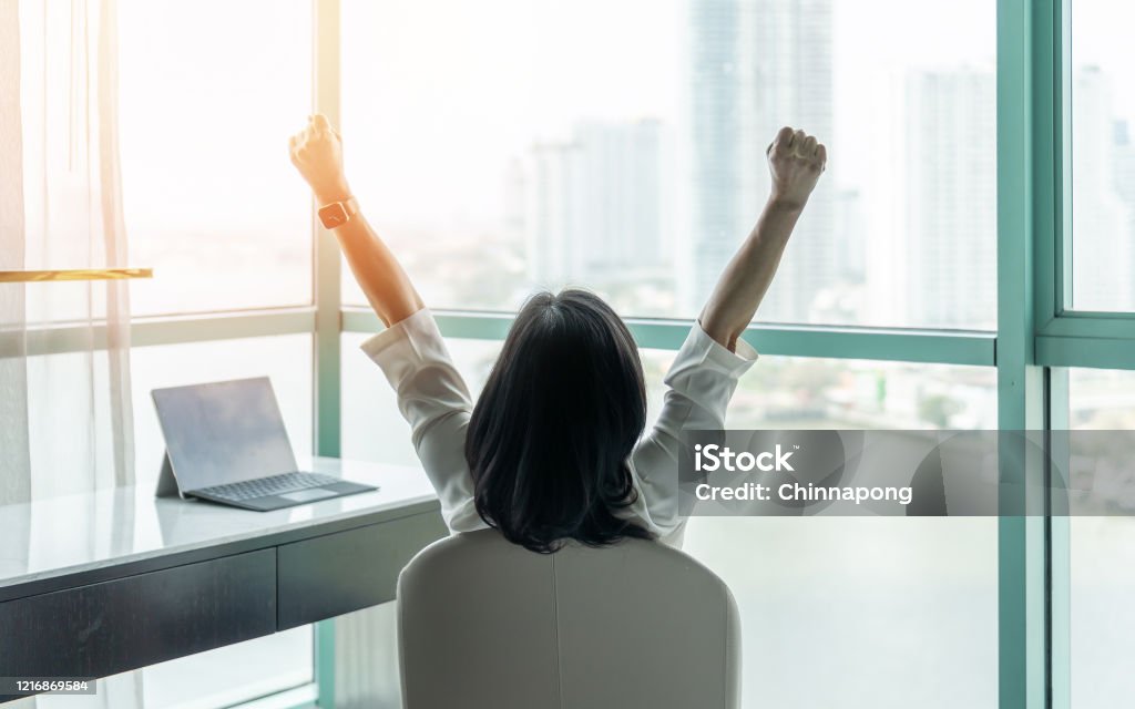 Business achievement concept with happy businesswoman relaxing in office or hotel room, resting and raising fists with ambition looking forward to city building urban scene through glass window Success Stock Photo
