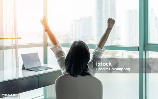 Business Achievement Concept With Happy Businesswoman Relaxing In Office Or Hotel Room Resting And Raising Fists With Ambition Looking Forward To City Building Urban Scene Through Glass Window Stock Photo - Download Image Now