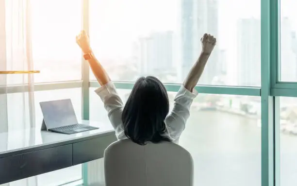 Photo of Business achievement concept with happy businesswoman relaxing in office or hotel room, resting and raising fists with ambition looking forward to city building urban scene through glass window