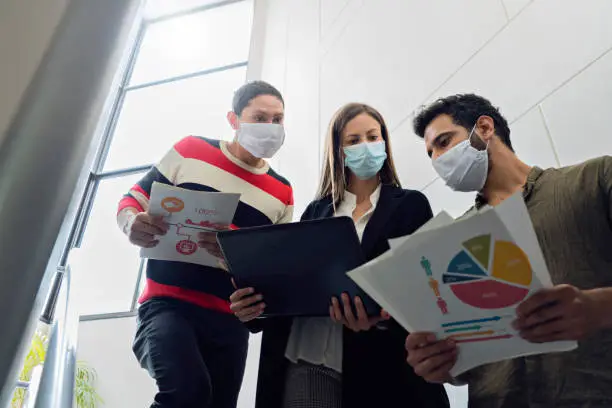 Photo of three coworkers are on the stairs of the company where they work with graphic documents and laptop all three wear masks