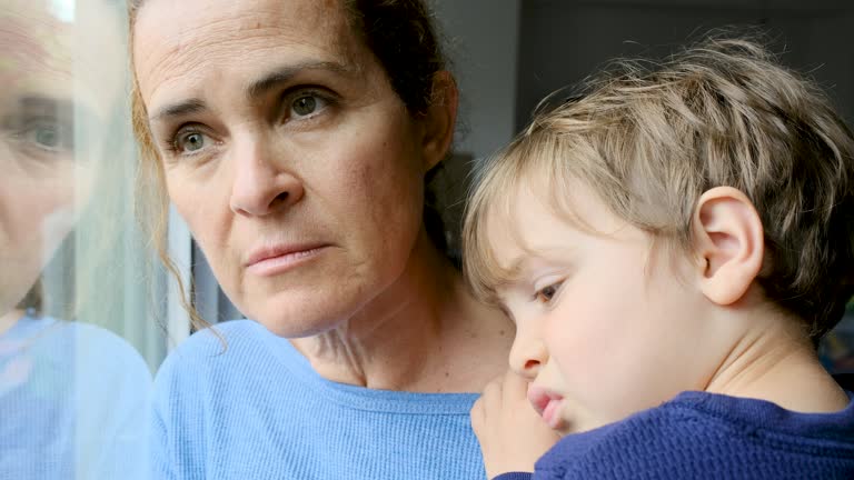 Mature woman posing with her son, very sad looking through window worried about Covid-19 lockdown