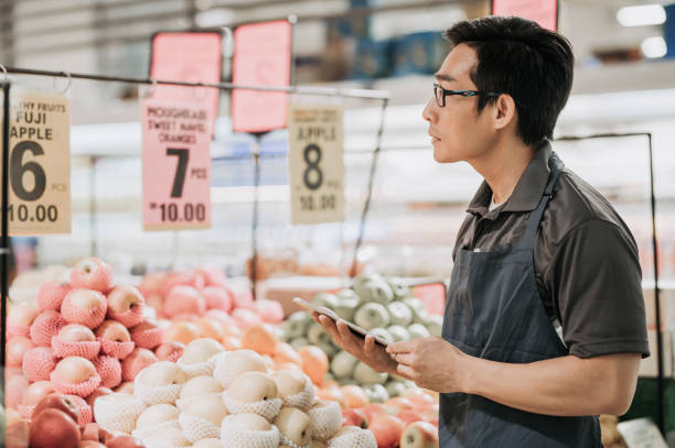 彼のデジタルタブレットでフルーツ店のデパートの前で在庫と値札をチェックアジアの中国の男性店アシスタント - supermarket sales clerk grocer apron ストックフォトと画像