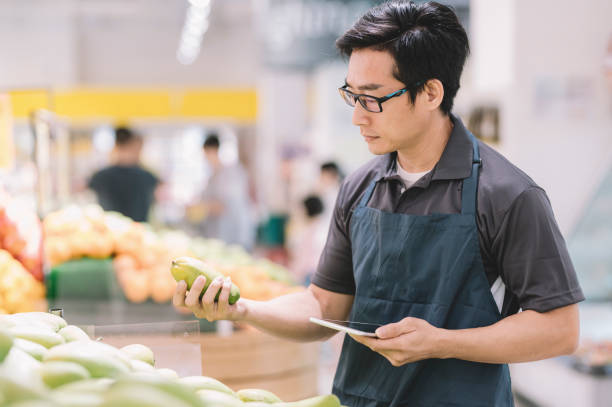 彼のデジタルタブレットでフルーツ店のデパートの前で在庫と値札をチェックアジアの中国の男性店アシスタント - supermarket sales clerk grocer apron ストックフォトと画像