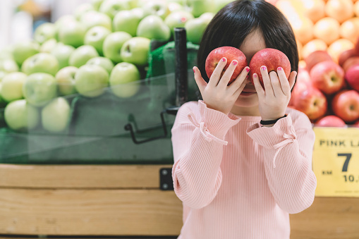 an asian chinese young girl holding 2 red apple in front of fruit store looking at camera