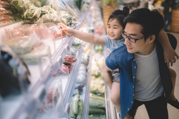 an asian chinese mid adult carrying his daughter at his back in supermarket choosing and buying from refrigerated section vegetables an asian chinese mid adult carrying his daughter at his back in supermarket buying tomatoes shopping asia stock pictures, royalty-free photos & images