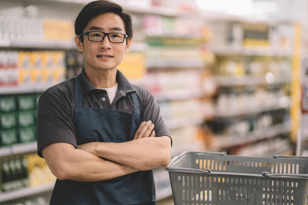 an asian chinese sales cashier standing at his cashier counter checkout place looking at camera smiling arm crossed an asian chinese sales cashier standing at his cashier counter checkout place looking at camera smiling arm crossed asian cashier stock pictures, royalty-free photos & images