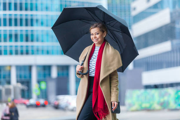 雨。美しいブルネットの女の子のアウトドアライフスタイルファッションの肖像画。街の通りを歩く。買い物に行く。スタイリッシュな白のフィットコート、赤いネックスカーフ、黒の傘の� - neckscarf ストックフォトと画像