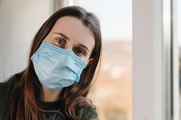 Close up portrait of brunette woman sit on windowsill with virus mask, thinks abot risk of epidemic disease, girl looks sadly at camera. Concept health and safety, N1H1 coronavirus, virus protection cerca del retrato de la mujer morena sentarse en el alféizar de la ventana con máscara de virus, piensa el riesgo de abot de enfermedad epidémica, chica mira tristemente a la cámara. concepto de salud y seguridad, coronavirus n1h1, protección antiviru