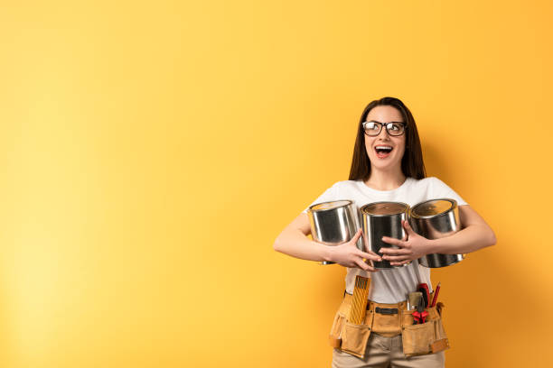 shocked repairwoman holding paint cans and looking away on yellow background shocked repairwoman holding paint cans and looking away on yellow background woman wearing tool belt stock pictures, royalty-free photos & images