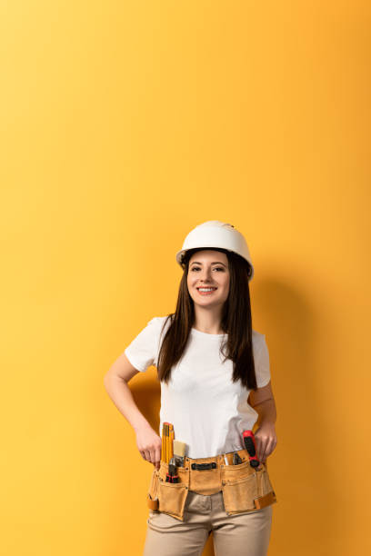 smiling handywoman in helmet looking at camera on yellow background smiling handywoman in helmet looking at camera on yellow background woman wearing tool belt stock pictures, royalty-free photos & images