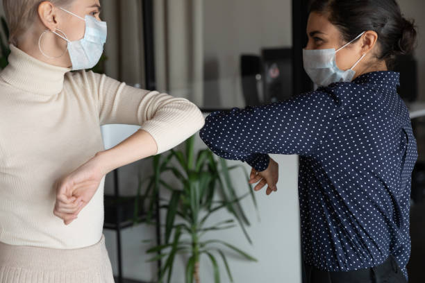 Healthy diverse colleagues in facial masks bumping elbows. Smiling young healthy mixed race female colleagues wearing facial medical masks greeting each other by bumping elbows gesture at workplace keeping social distance, preventing spreading covid19 virus. elbow photos stock pictures, royalty-free photos & images