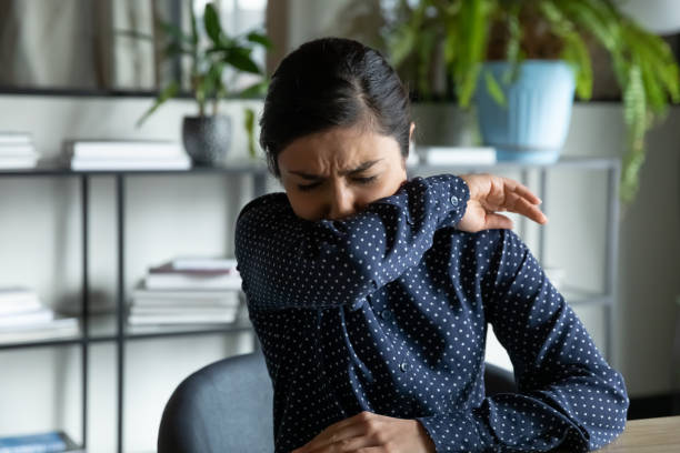 Unhealthy young woman feeling unwell at workplace, covid 19 symptoms. Millennial indian ethnic girl sitting in office, coughing in elbow, right illness behavior for not spreading virus infection. Unhealthy young woman feeling unwell at workplace, covid 19 symptoms. sneezing photos stock pictures, royalty-free photos & images