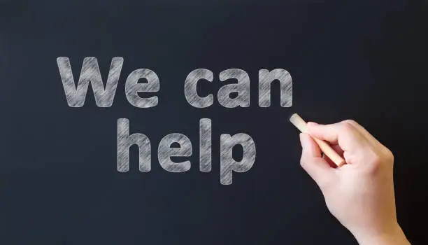 Photo of We can help - chalk inscription on a black chalk board. Hand with chalk writes text